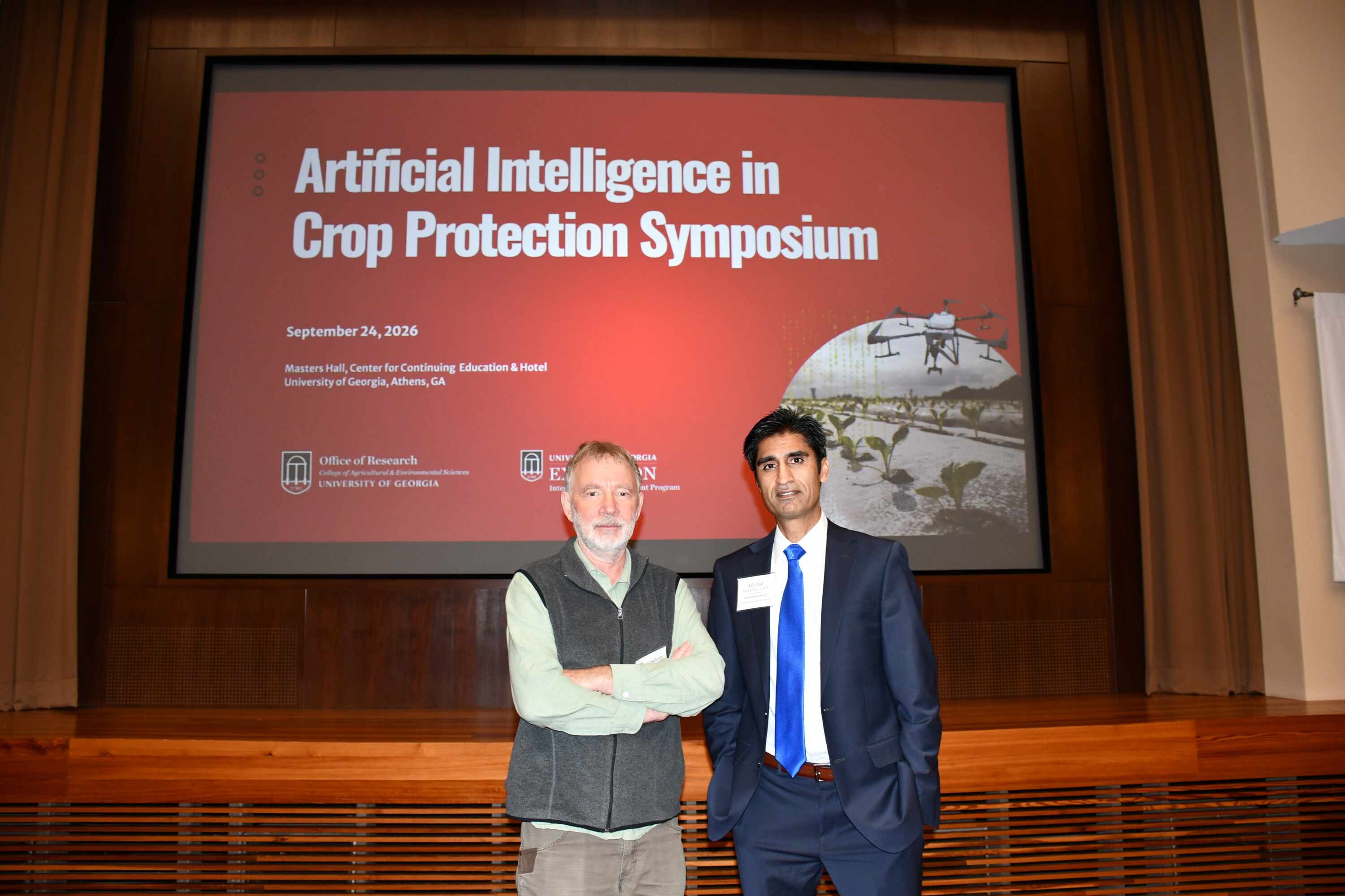 A man in a gray vest and a blue suit pose in front of a slide that says "Artificial Intelligence in Crop Protection Symposium."